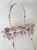 Cartera de cuero de tamaño medio con estructura rígida. Presenta una tira decorativa con tachas y hebillas metálicas en los extremos, además de lazos laterales. Cuenta con doble asa para llevar al hombro y cierre superior.