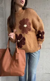 Sweater de punto grueso color camel, con diseño de flores grandes aplicadas en tonos marrones y centro crema. Presenta cuello redondo alto, mangas largas con hombros caídos y terminaciones acanaladas.