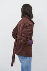 Chaqueta de gamuza en tono marrón oscuro, con cuello alto, cierre frontal con cremallera y botones a presión, bolsillos frontales con solapa y cinturón ajustable en la cintura.