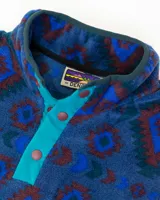Buzo de polar fleece con diseño estampado geométrico en tonos azules, bordó y verde. Presenta cuello alto con cierre de botones a presión, bolsillo frontal con solapa en contraste y puños elásticos.