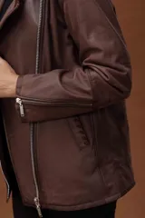 Chaqueta estilo motoquera de cuero ovino color marrón, con diseño oversize. Presenta detalles artesanales de pasamanería en cuello, solapas y bolsillos, cierre frontal cruzado y cierres metálicos en los puños.