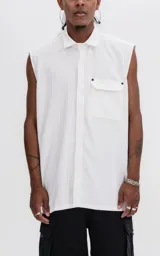 Camisa sin mangas de corte oversize, color blanco, con cuello clásico, cierre frontal oculto y bolsillo tipo cargo en el pecho con solapa y remaches metálicos.