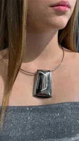 Gargantilla tipo choker rígida de metal plateado con un dije central grande de forma rectangular y acabado espejado.