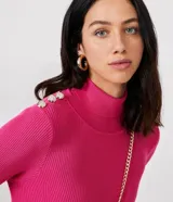 Vestido corto de punto acanalado negro, con cuello alto, mangas largas y falda acampanada. Presenta detalle de botones dorados en los hombros.