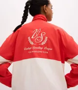 Campera deportiva de cuello alto con diseño color blocking en rojo y blanco. Presenta cierre frontal parcial, mangas largas con puños elásticos y bordado decorativo en el pecho. Confeccionada en poliamida con tecnología dry para mayor transpirabilidad.