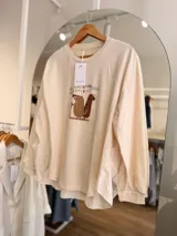 Buzo liviano de media estación color beige, confeccionado en 100% algodón, con estampa frontal ilustrada de osos y texto. Presenta calce oversize, mangas largas y diseño de largo asimétrico con espalda más larga.