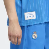 Remera de fútbol Adidas del Real Madrid, modelo Terrace Icons. Diseño celeste con finas rayas verticales blancas, cuello redondo blanco, mangas cortas con tres tiras blancas en los hombros, logo de Adidas y escudo del Real Madrid bordados en el pecho. Inspirada en la moda del fútbol de los años 80.