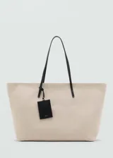 Bolso tipo shopper de gran tamaño confeccionado en tejido de canvas color beige. Cuenta con dos asas de hombro largas en contraste color negro, cierre de cremallera y un charm colgante decorativo en color negro.
