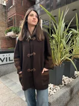 Saco capa de lana color marrón oscuro, con diseño de punto fino y cierre frontal con tres botones de asta y trabillas de cuero. Cuenta con mangas anchas con detalles texturados en los codos, bolsillos frontales aplicados con ribete de pana y silueta holgada.