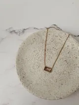 Collar de cadena fina dorada con un dije rectangular que contiene una piedra de cristal en tono rosado.