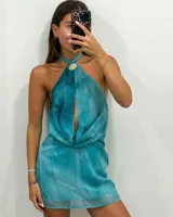 Vestido corto color turquesa con efecto tie-dye, escote halter que se anuda al cuello con un dije redondo dorado y abertura frontal con drapeado en el busto.