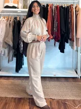 Blusa de tela liviana con diseño de rayas verticales en tonos beige y crema. Presenta cuello alto con detalle de fruncido tipo smock, mangas largas abullonadas con puños elásticos y volados en los extremos.