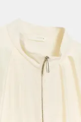 Chaqueta bomber de cuello subido y manga larga, con acabado satinado en color beige. Presenta pliegues frontales, bolsillos laterales ocultos en costura y cierres elásticos en puños y cintura. Cierre frontal con cremallera.