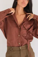 Camisa blanca de manga larga, con botones de guampa y un detalle de roseta o flor a tono en la parte delantera. El calce es holgado con la espalda ligeramente más larga.