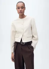 Chaqueta corta de cuello redondo y manga larga, con cierre frontal de botones y dos bolsillos laterales con solapa. Presenta un diseño recto y estructurado.