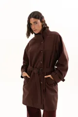 Trench color chocolate con cuello alto, cierre frontal con botones y cinturón ajustable en la cintura. Presenta mangas largas con puños ajustables mediante tiras y hebillas, y bolsillos frontales.
