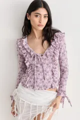 Blusa de encaje color lila con estampado floral, escote en V pronunciado con volados y lazo frontal. Posee mangas largas con detalles de volados en hombros y puños.