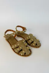 Sandalias planas de gamuza color tostado, con múltiples tiras decoradas con tachas metálicas doradas semiesféricas. Cuenta con una tira ajustable en el tobillo con hebilla metálica.