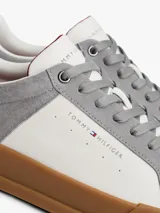 Zapatillas de tenis Tommy Hilfiger de corte bajo, confeccionadas en piel nobuk con paneles de texturas mixtas en blanco y gris. Presentan suela de goma color caramelo, cierre con cordones y detalles distintivos de la marca en la lengüeta, lateral y talón.