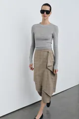 Falda midi de tiro medio con estampado de cuadros en tonos beige y gris. Presenta un diseño cruzado con un volante decorativo en el delantero y cierre con botón.