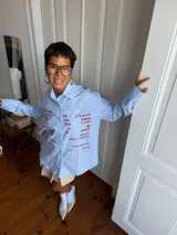 Camisa oversize de corte recto, confeccionada en tela de algodón a rayas verticales celestes y blancas. Presenta cuello clásico, cierre frontal con botones y mangas largas. El diseño destaca por una estampa frontal con palabras relacionadas al amor en diversos idiomas en color bordó.