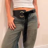 Cinto ancho de cuero marrón con diseño asimétrico, que presenta un aro metálico dorado como elemento central y una tira de gamuza larga que se anuda para el cierre.