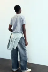 Camiseta gris de cuello redondo y manga corta, con un estampado en relieve que dice "NEW YORK CITY" y "BROOKLYN". El tejido tiene un sutil brillo.