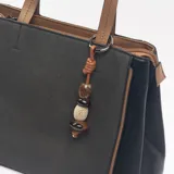 Cartera tipo tote de diseño estructurado, combinando paneles de tela gris oscuro con laterales y detalles en cuero sintético negro y marrón. Incluye doble asa superior, correa larga ajustable y extraíble, y un dije decorativo con cuentas de madera y piedra.