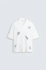 Camisa de manga corta confeccionada en mezcla de algodón y lino, color blanco. Presenta un cuello tipo bowling, calce regular y cierre frontal con botones. Destaca por sus bordados lineales en contraste de color negro con motivos de frutas y hojas distribuidos por toda la prenda.