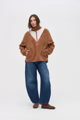 Buzo de sherpa color marrón con diseño de medio cierre frontal. Presenta un panel superior en forma de V en color beige y detalles de vivos en color fucsia en el cierre, cuello y bolsillos laterales. Cuenta con cuello alto y bolsillos frontales.