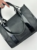 Bolso tote negro de cuero con detalles de charol, doble asa corta y correa bandolera de tela con letras.
