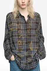 Camisa de corte oversize con cuello camisero, cierre frontal de botones y mangas largas con puños abotonados. Presenta un diseño estampado que combina un patrón de cuadros con motivos decorativos en tonos marrones, azules y amarillos.
