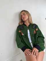 Campera oversize verde con apliques de cuero negro en los hombros y detalles de cuero color cobre en el frente. Tiene cuello redondo, cierre con botones a presión y puños negros.