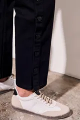 Pantalón de jean negro, de corte recto y tiro medio. Presenta bolsillos delanteros y traseros, y una hilera de botones metálicos a lo largo de la costura lateral.