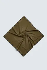 Pañuelo cuadrado de algodón color verde oliva con un patrón de pequeñas cruces blancas distribuidas por toda la superficie. El borde presenta un ribete con un patrón de cuadros en tonos oscuros (negro, azul marino) y líneas finas blancas.