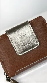 Billetera compacta de cuero texturizado color marrón, con solapa frontal en cuero metalizado dorado que presenta un grabado de búho. Incluye cierre lateral y correa de muñeca a juego.