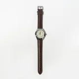 Reloj analógico con caja plateada, esfera blanca con números arábigos y correa marrón de cuero.