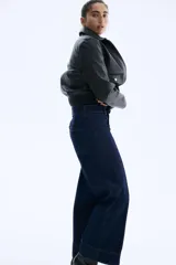 Jean de tiro alto en denim azul oscuro con pierna amplia y corte tobillero. Presenta un diseño de cinco bolsillos, cierre frontal con botón y costuras decorativas verticales en el frente que simulan un pliegue.