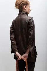 Blazer de cuero marrón con cierre frontal, cuello alto, múltiples bolsillos con hebillas y detalle de triángulo de cuero en el pecho.