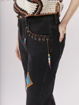 Pantalón de jean tiro alto tipo mom-fit, con parches de colores en forma geométrica, detalles de tientos de cuero trenzados en la cintura y adornos de canutillos en la parte frontal.