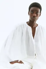 Blusa de ramio color blanco, con cuello redondo y cierre frontal de botones nacarados. Presenta un diseño de corte holgado con canesú fruncido en hombros y espalda, y mangas largas amplias que aportan volumen a la silueta.