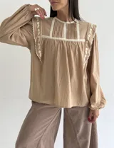 Blusa color beige de algodón con cuello redondo, mangas largas con puños elásticos y detalles de puntilla y bordados en el frente y hombros.