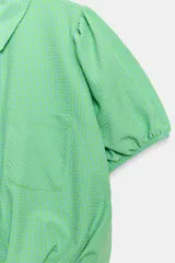 Camisa corta de cuello solapa y manga corta abullonada acabada en elástico, color verde con estampado de cuadros vichy. Cuenta con un bolsillo de plastrón en el delantero, bajo con elástico y cierre frontal con botones.