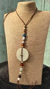 Collar largo con tiras de cuero marrón, decorado con cuentas de diferentes materiales y un dije circular central de nácar.