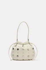 Bolso estilo saca de cuero color marfil, con diseño fruncido en la parte superior, tiras ajustables y tachas metálicas plateadas distribuidas por toda la superficie. Cuenta con una manija superior.