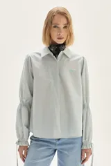 Camisa a rayas verticales de calce recto, con cuello clásico y mangas abullonadas con detalles de frunces y lazos en los puños. Presenta un pequeño bordado en el pecho.