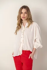 Camisa color crema de corte holgado, con cuello mao, cierre frontal con botones y volantes, mangas largas con puños elásticos y bordados en hombros y puños.