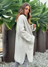 Sacón de punto color crudo con ribetes marrones, de corte largo y holgado, con cuello redondo, mangas largas y dos bolsillos frontales.