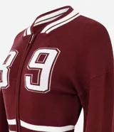 Cardigan de punto estilo college color bordó, con cierre frontal de doble cremallera, mangas largas y detalles de rayas blancas en cuello, puños y ruedo. Presenta una estampa numérica blanca en el frente.