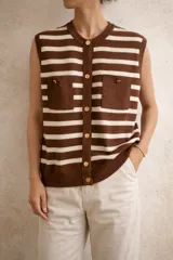 Chaleco tejido de punto sin mangas, con diseño de rayas horizontales en color marrón y blanco. Presenta cierre frontal con botones dorados, cuello redondo y dos bolsillos frontales tipo parche.